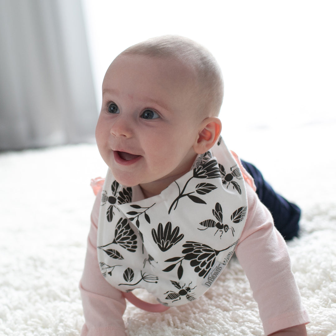 A baby wearing a Dr. Brown’s™ Bandana Bib with Removable Teether crawls on a soft, white carpet. The baby looks up with a joyful expression in a softly lit room.