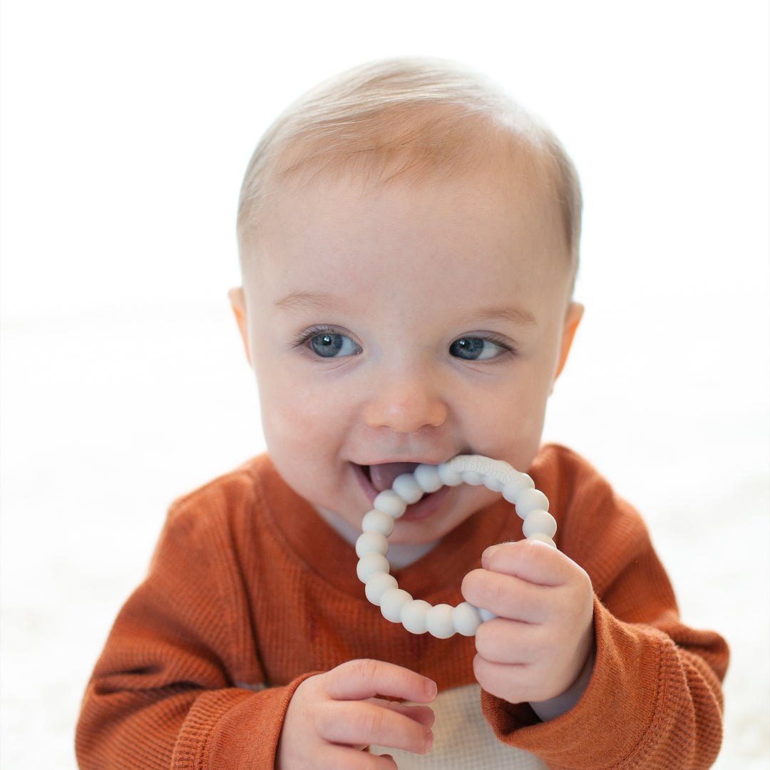 A baby with light hair and blue eyes is dressed in a rust-colored outfit, holding Dr. Brown's Dr. Brown’s™ Flexees™ Beaded Teether Rings. The child playfully nibbles on it to soothe teething pain, smiling gently in the softly lit setting, finding comfort as they self-soothe.