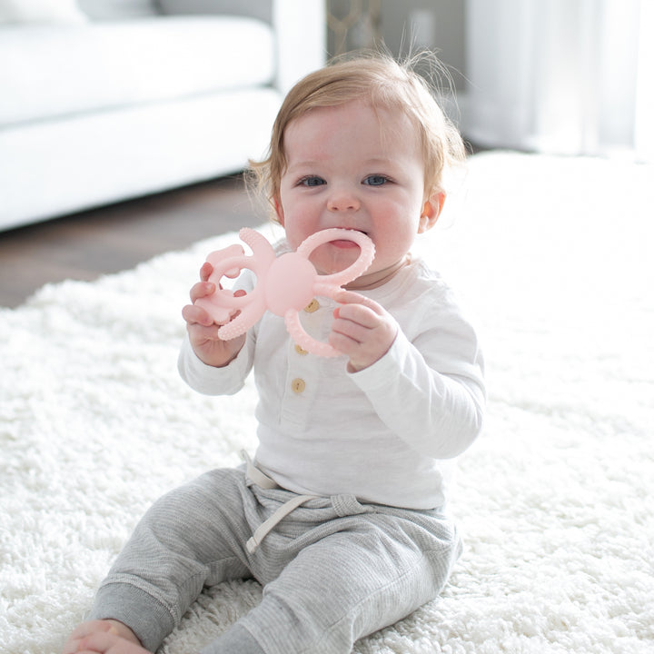 A baby sits on a white carpet, holding a Dr. Brown's™ Flexees™ Bunny Teether in pink for teething relief. The child is dressed in a white long-sleeve shirt and gray sweatpants. Sunlight filters through the window behind, casting a soft glow over this serene moment.