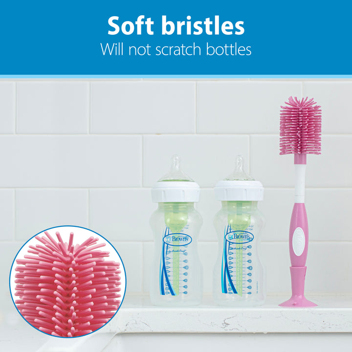 A Dr. Brown’s™ Soft Touch Bottle Brush in pink, featuring soft bristles and a durable design, rests on a kitchen counter with two baby bottles beside it. A close-up inset highlights the bristles. Text reads, "Scratch-free scrubbing with soft bristles will not scratch bottles," emphasizing its gentle cleaning capabilities.