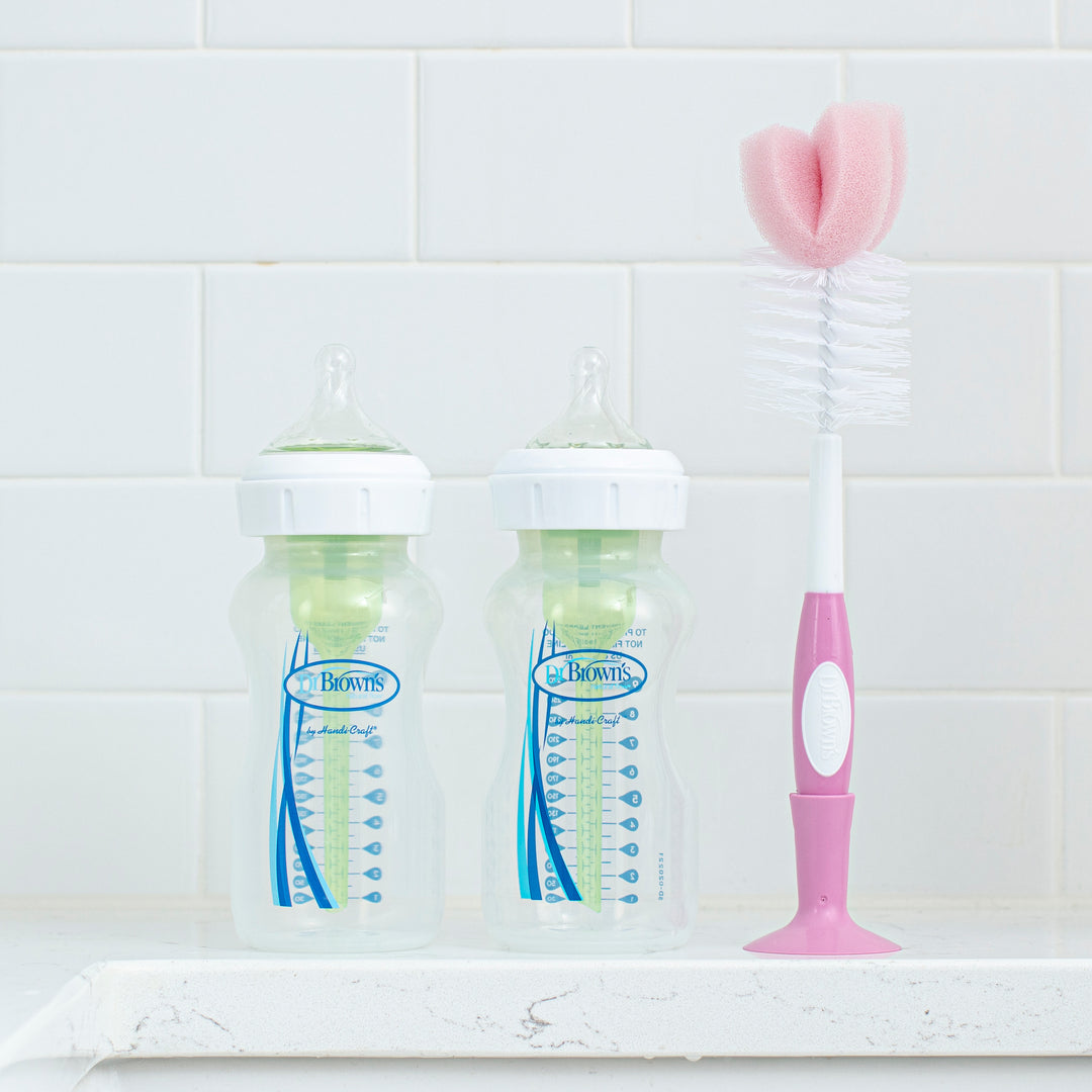 Two clear baby bottles with green inserts and blue markings stand on a white counter against a tiled wall. Beside them, the Dr. Brown’s Natural Flow® Baby Bottle Brush—a popular choice among parents—features a pink and white handle and sponge tip, positioned upright and ready to also serve as an effective nipple cleaner.