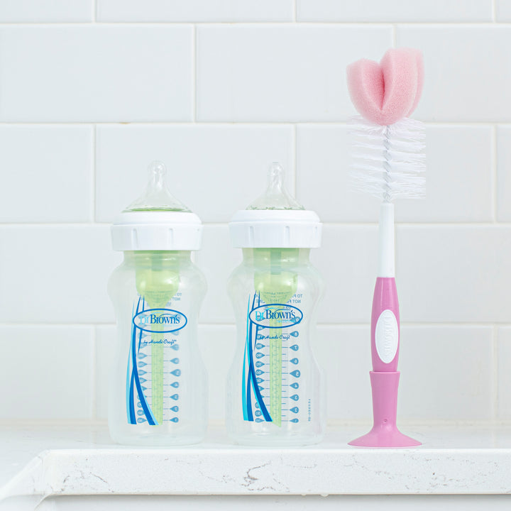 Two clear baby bottles with green inserts and blue markings stand on a white counter against a tiled wall. Beside them, the Dr. Brown’s Natural Flow® Baby Bottle Brush—a popular choice among parents—features a pink and white handle and sponge tip, positioned upright and ready to also serve as an effective nipple cleaner.