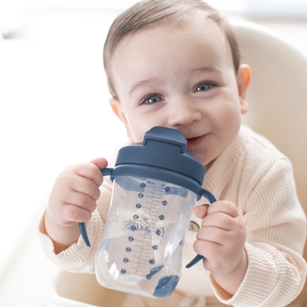 A smiling baby in a beige outfit sits in a high chair, holding their Dr. Brown's™ Milestones™ Baby’s First Straw Cup. The cup has a blue handle and features a clear design with measurement markings to assist with the bottle-to-cup transition. Its weighted straw allows for easy sipping.