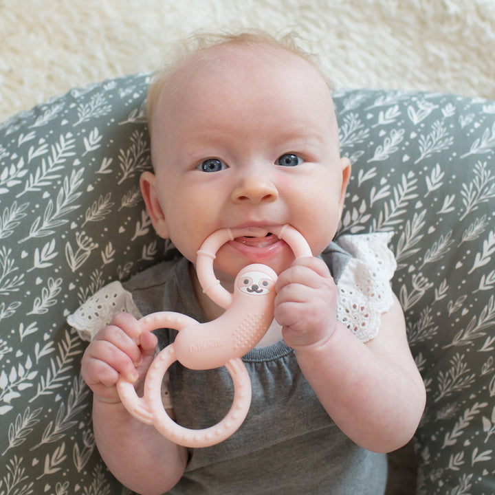 A baby is lying on a patterned cushion, happily chewing on a pink Dr. Brown’s™ Flexees™ Sloth Teether. The little one looks content in a gray outfit with lace sleeves, snugly holding their favorite teething aid from Dr. Brown's.