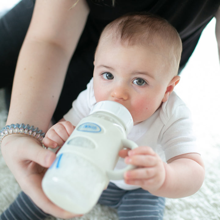 A baby dressed in a white shirt and striped pants sits indoors, sipping milk from Dr. Brown’s® Milestones™ Wide-Neck Bottle with silicone handles, held by an adult. The baby looks upward while developing independent drinking skills on a soft surface.