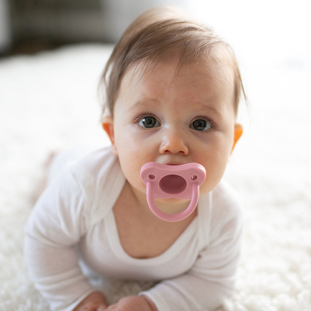 A baby with brown hair lies on a white fuzzy carpet, wearing a white onesie and gently holding Dr. Brown’s® HappyPaci™ 100% Silicone One-Piece Pacifier. The baby gazes directly at the camera, exuding tranquility.