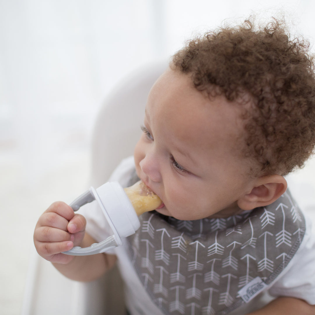 A baby with curly hair sits in a high chair, holding Dr. Brown's™ Fresh Firsts™ Silicone Feeder filled with an edible treat. Wearing a patterned bib adorned with arrow designs, the baby looks to the side while biting the toy.