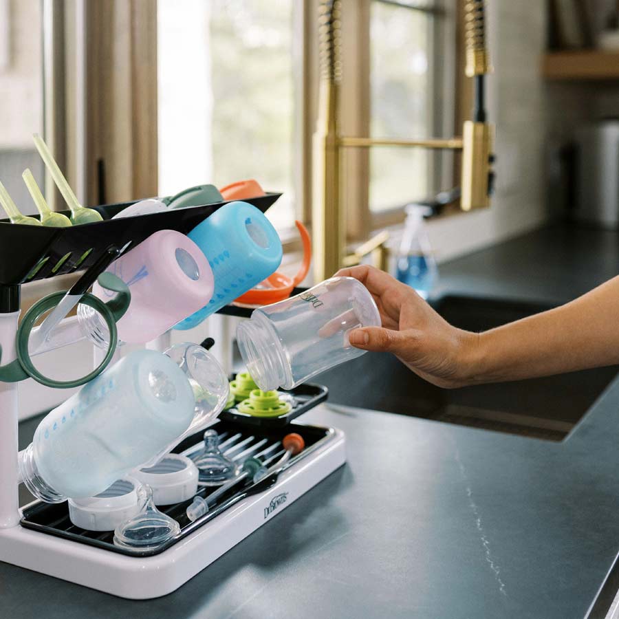 A person places a clear baby bottle onto Dr. Brown's™ Drying Tower, which accommodates an assortment of colorful bottles, lids, and other small items. This countertop space-saving solution is situated on the kitchen counter beside a sink with gold faucets.