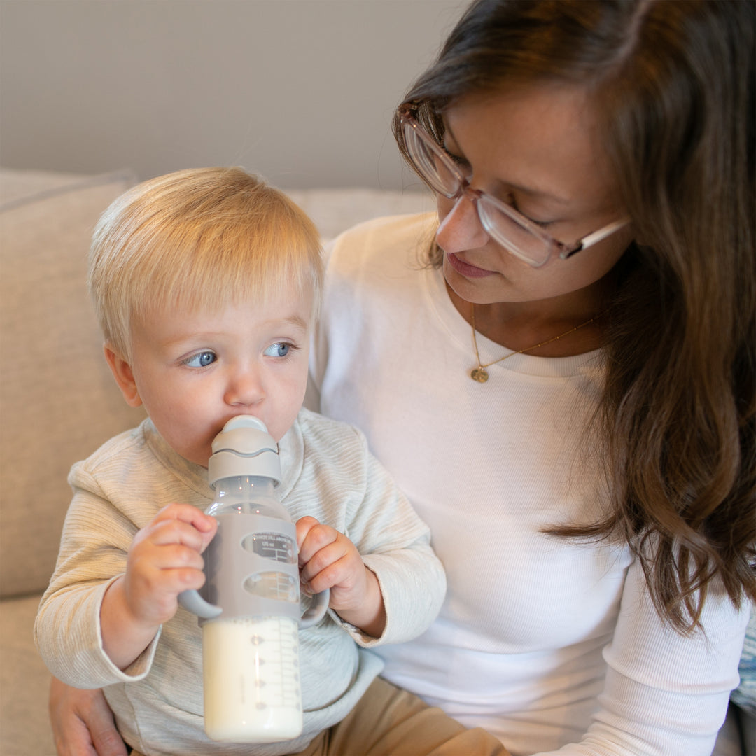 A bespectacled woman holds a young child sipping from a Dr. Brown's™ Milestones™ Narrow Sippy Straw Bottle with Silicone Handles, 8 oz/250 mL, as they sit on a couch in light-colored attire. The peaceful and nurturing atmosphere is enhanced by the bottle’s spill-proof lid, allowing the child to develop independent drinking skills without creating any mess.