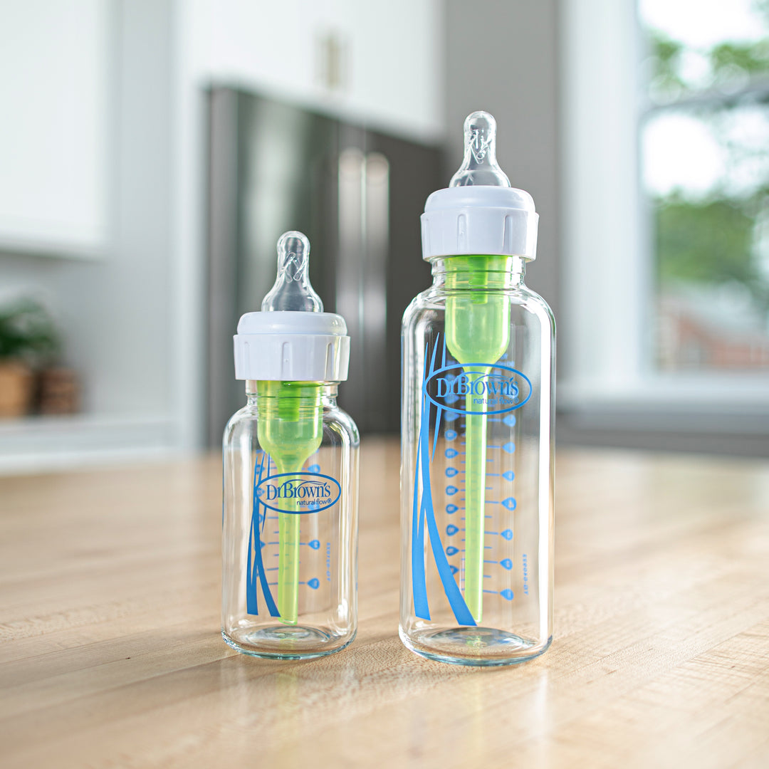 Two Dr. Brown’s Natural Flow® Anti-Colic Options+™ Narrow Glass Baby Bottles, featuring blue and green accents, stand on a wooden kitchen counter. Designed to reduce colic, they come with white caps and Level 1 Slow Flow silicone nipples, complete with measurement markings on the sides. Bright window light illuminates the background, ensuring natural flow visibility.