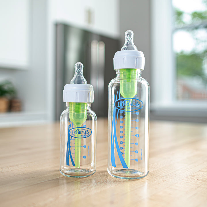 Two Dr. Brown’s Natural Flow® Anti-Colic Options+™ Narrow Glass Baby Bottles, featuring blue and green accents, stand on a wooden kitchen counter. Designed to reduce colic, they come with white caps and Level 1 Slow Flow silicone nipples, complete with measurement markings on the sides. Bright window light illuminates the background, ensuring natural flow visibility.