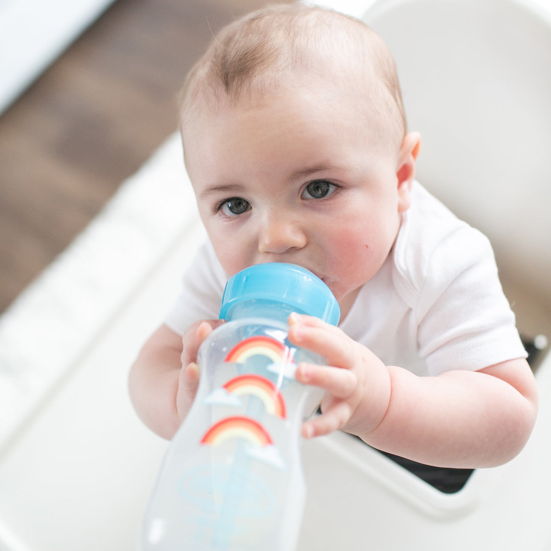 A baby in a white onesie sits in a high chair, sipping from the Dr. Brown’s Natural Flow® Anti-Colic Options+™ Wide-Neck Sippy Bottle Starter Kit, 9oz/270mL, which features a colorful rainbow design and a soft silicone spout. The indoor setting with a light background ensures comfort and an anti-colic experience for the little one.