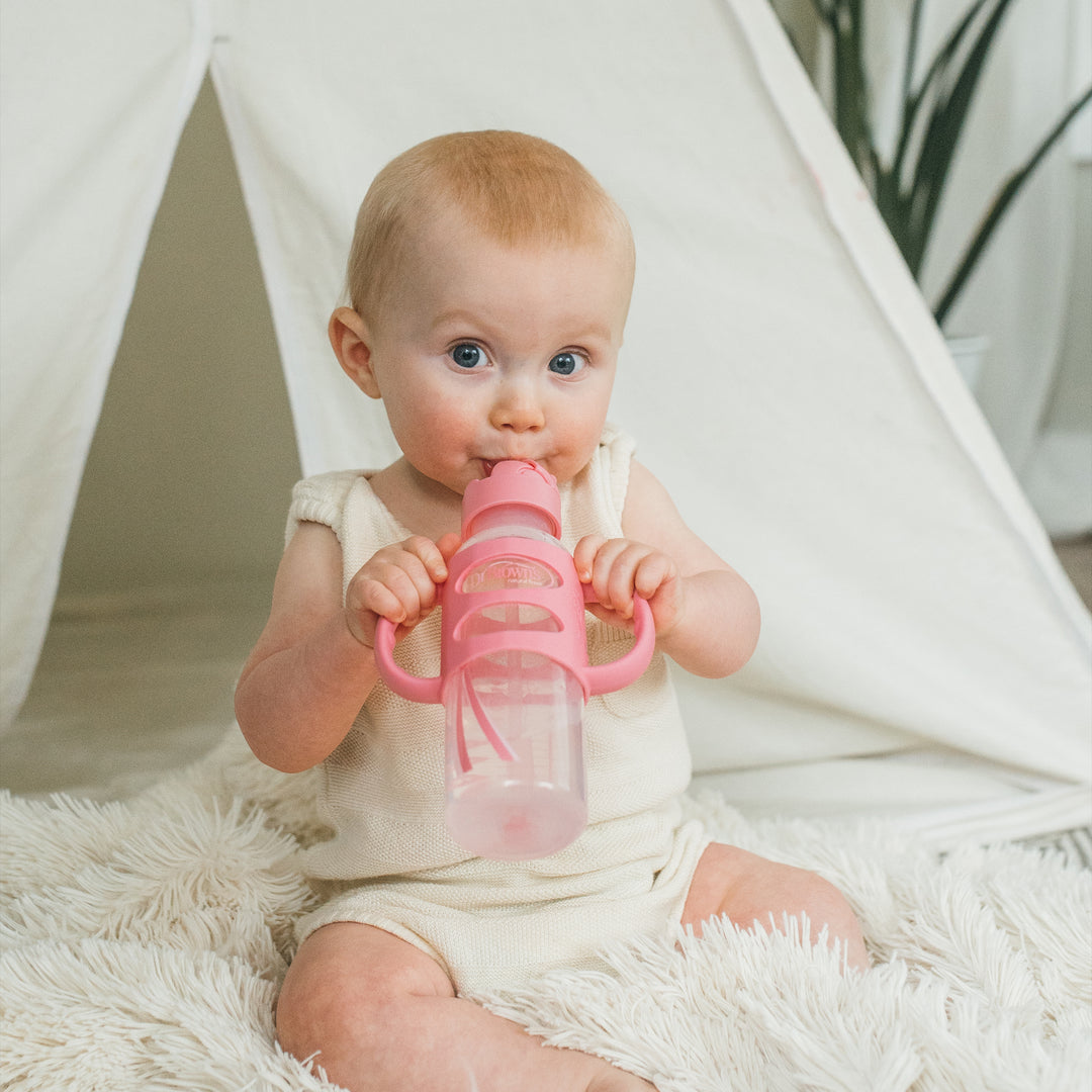 A baby with short, light hair is sitting on a fluffy rug, dressed in a cream outfit and drinking from Dr. Brown’s™ Milestones™ Narrow Sippy Straw Bottle with Silicone Handles, 8 oz/250 mL, in pink. Behind the baby, there is a white tent and a green plant. This bottle features a silicone weighted straw to foster independent drinking skills.