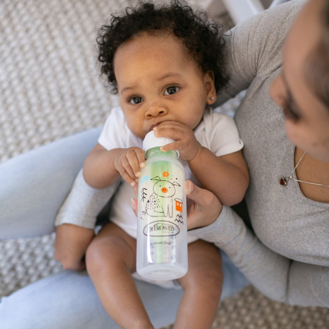 A baby with curly hair is seated on an adult's lap, sipping from a Dr. Brown’s Natural Flow® Anti-Colic Options+™ Narrow Baby Bottle, featuring vibrant cartoon designs. The adult, wearing a gray top and jeans, cradles the baby tenderly in a cozy environment to ensure each sip helps prevent colic discomfort.