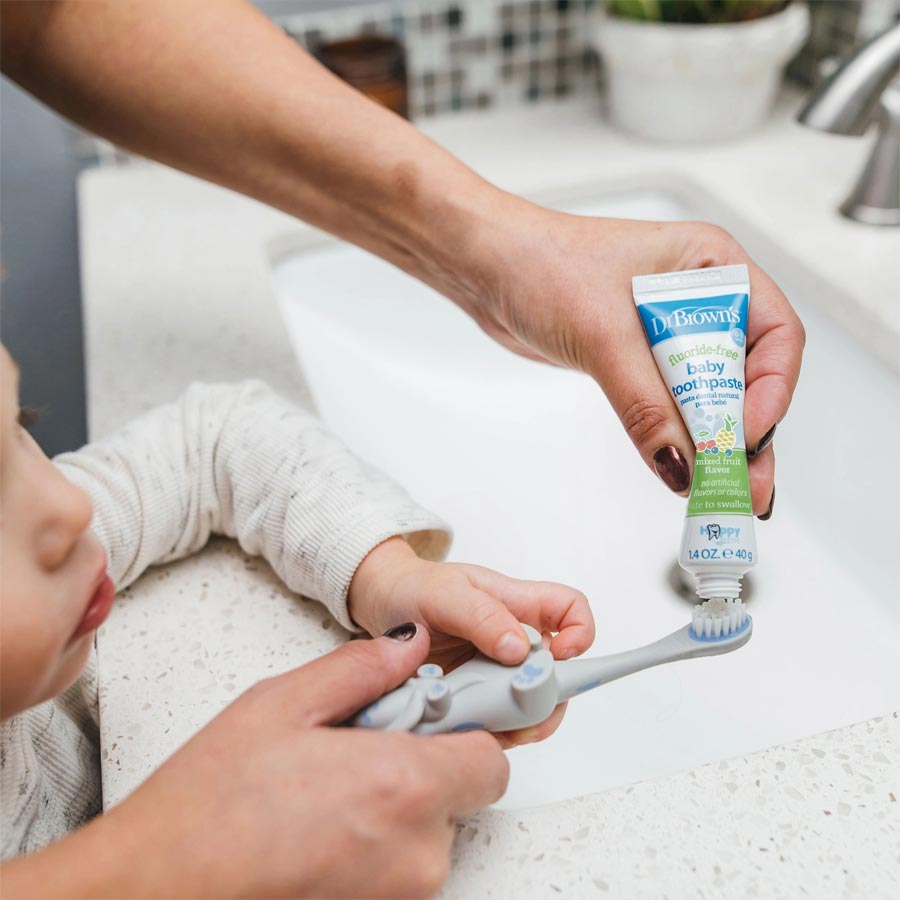 An adult is gently squeezing Dr. Brown's™ Fluoride-Free Baby Toothpaste [Mixed Fruit] from a tube onto a toothbrush held by a child. The child, dressed in a white sweater, leans over the bathroom sink. In this well-lit bathroom complete with countertop and faucet, early oral hygiene habits are being nurtured.