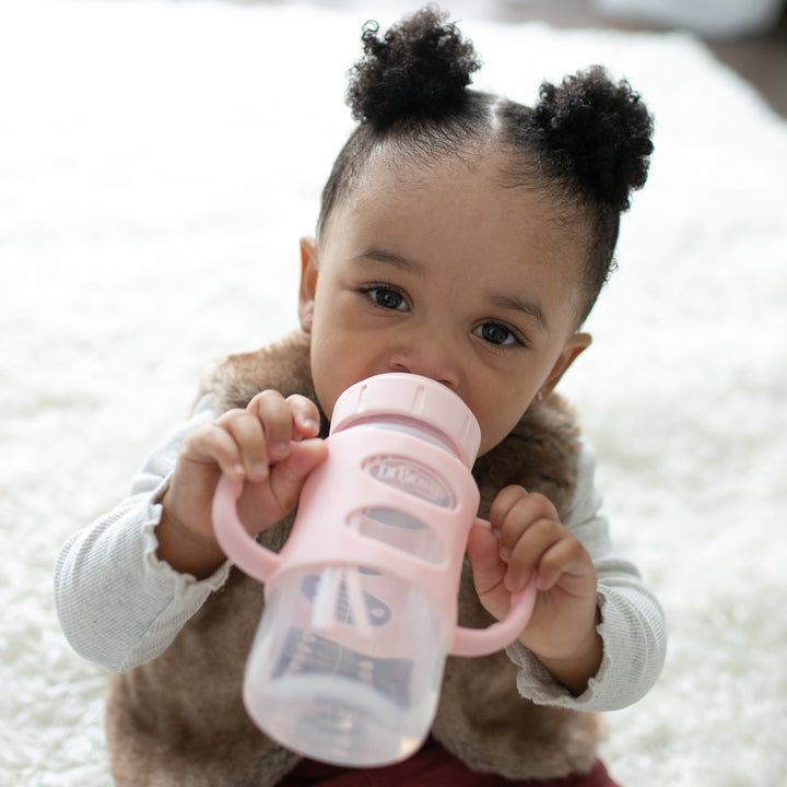 A toddler with two puffy hair buns is drinking from a Dr. Brown's® Milestones™ Wide-Neck Sippy Spout Bottle with Silicone Handles, 9 oz/270 mL. They are wearing a brown vest over a white shirt and are seated on a fluffy, white surface, practicing transition drinking skills.