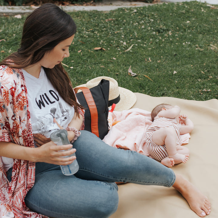 A woman relishes her mom time on a blanket in the park, holding a water bottle. Next to her, a baby lies on its back with hats and Dr. Brown’s Breast Pump Carryall Tote Bag nearby. The woman is comfortably dressed in jeans and a patterned shawl.