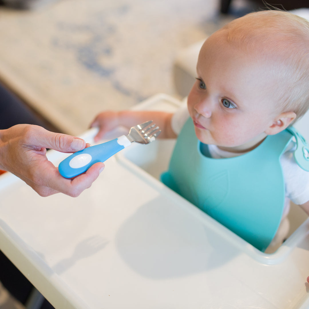 A baby in a highchair, wearing a turquoise bib, curiously eyes the Dr. Brown’s™ Designed to Nourish™ Soft-Grip Fork with a blue handle, held towards them by a hand. The baby, with light hair and seated at a white table, is surrounded by soft indoor lighting.