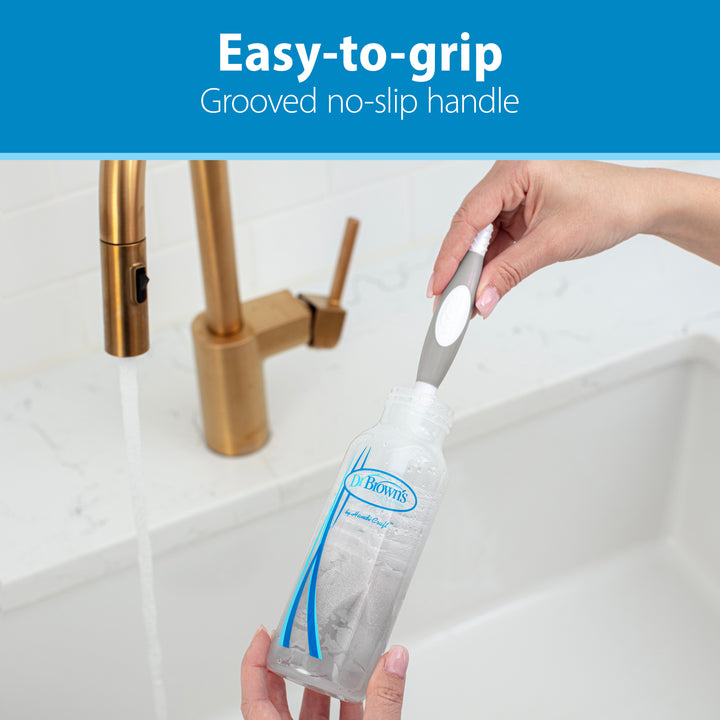 A person is shown cleaning a baby bottle using the highly popular Dr. Brown’s Natural Flow® Baby Bottle Brush under a running faucet, showcasing its "Easy-to-grip Grooved no-slip handle." The bottle bears the label "Dr. Brown's," and the sink and faucet have a gold-toned finish.