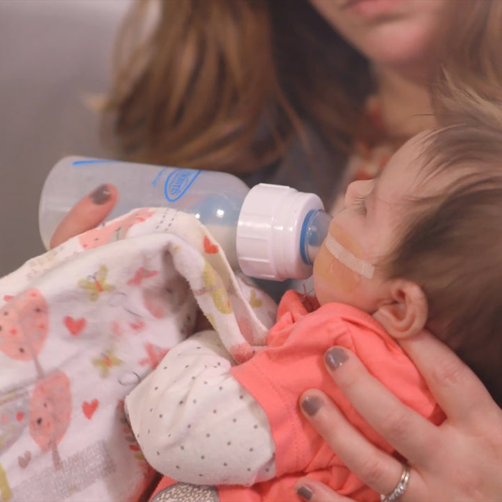 A person is holding a baby using the Dr. Brown’s® Specialty Feeding System, addressing oral feeding issues. The little one, with a small tube attached to their face, is nestled on a patterned blanket. The caregiver wears an orange shirt and patterned sleeves, demonstrating care and comfort in every detail.