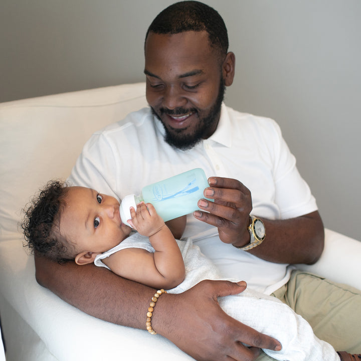 A bearded man in a white shirt is seated in a chair, smiling as he feeds a baby using Dr. Brown's Natural Flow® Options+™ Wide-Neck Glass Bottle. The bottle is complemented by silicone sleeves, providing protection, while the baby rests comfortably on his arm, dressed in a light-colored outfit. The moment captures warmth and tenderness.