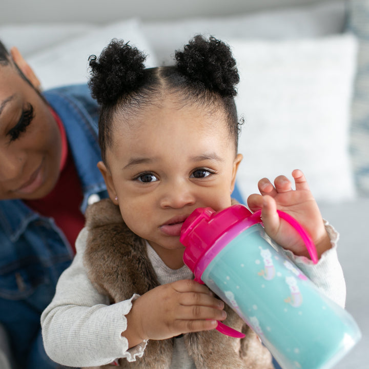 A toddler with two pigtails clutches a Dr. Brown's Milestones™ Insulated Straw Cup, resting against an adult's arm. The adult, partially visible on the left, wears a denim jacket. They're in a cozy indoor setting.