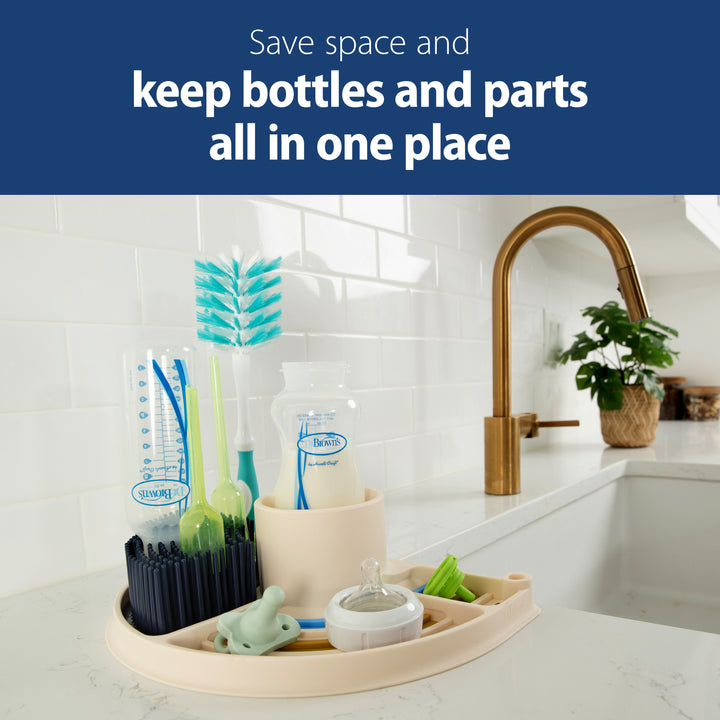 On a kitchen countertop, you'll find Dr. Brown’s™ 2-in-1 Dry & Prep Silicone Mat displaying a beige bottle drying rack with baby bottle parts, a cleaning brush, and a pacifier. A sleek modern faucet and potted plant complete the scene. Text reads: "Save space and keep bottles and parts all in one place.