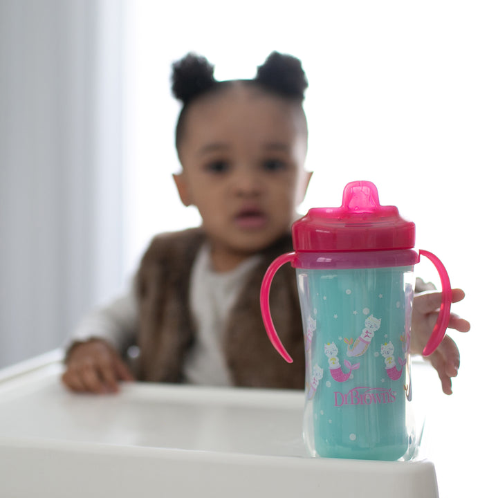 A toddler with hair in two buns sits in a high chair, wearing a white shirt and brown vest. In the foreground is one of the Dr. Brown’s Milestones™ Insulated Hard Spout Sippy Cups, from a 2-pack by Dr. Brown's, featuring a pink handle and blue center adorned with cartoon designs, known for its bite-resistant durability.