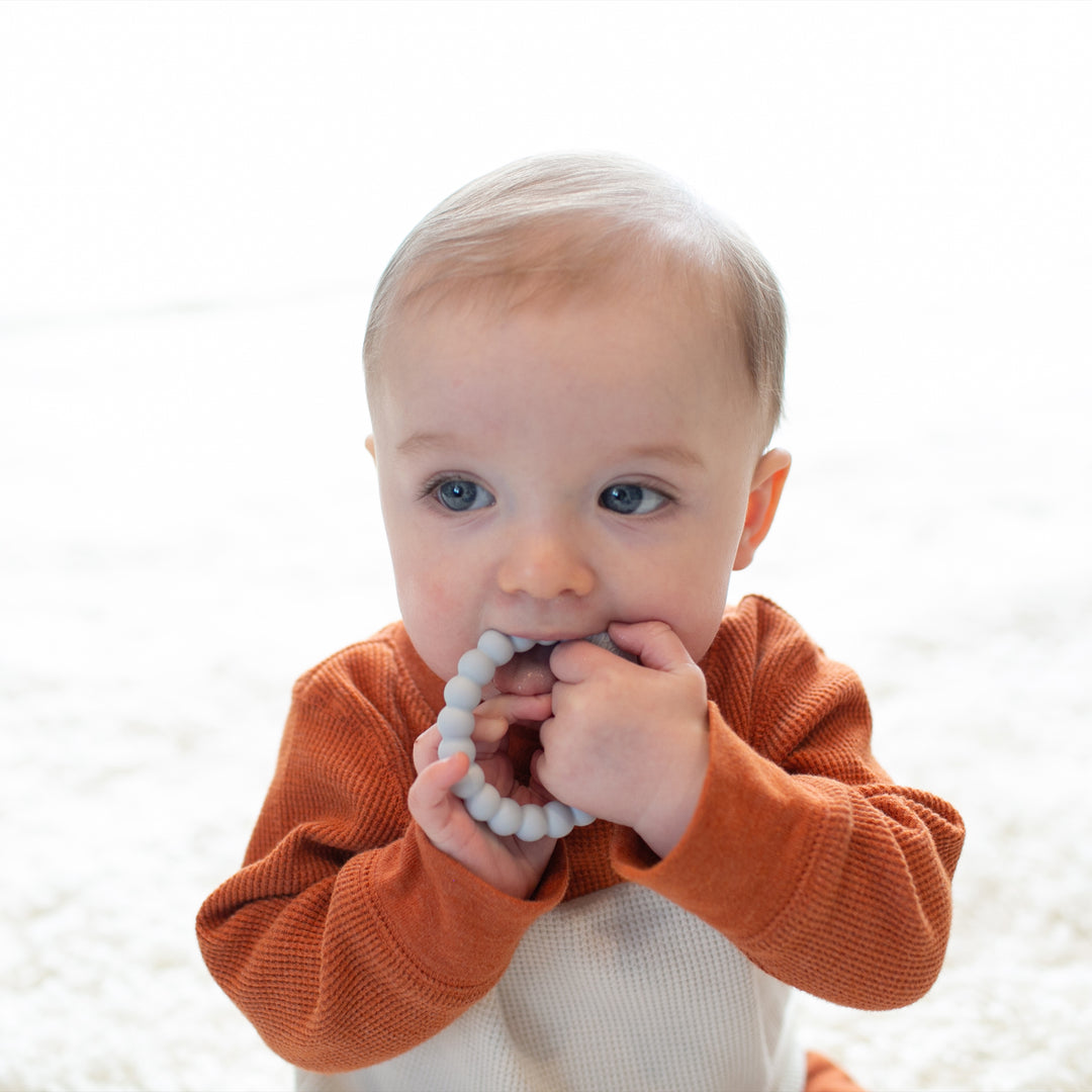 A baby with light hair in a rust-colored sweater sits on a soft, light-colored surface, chewing on Dr. Brown's™ Flexees™ Beaded Teether Rings. The little one grasps them with both hands, engaged and gazing slightly to the right—an adorable scene of self-soothe baby exploration.