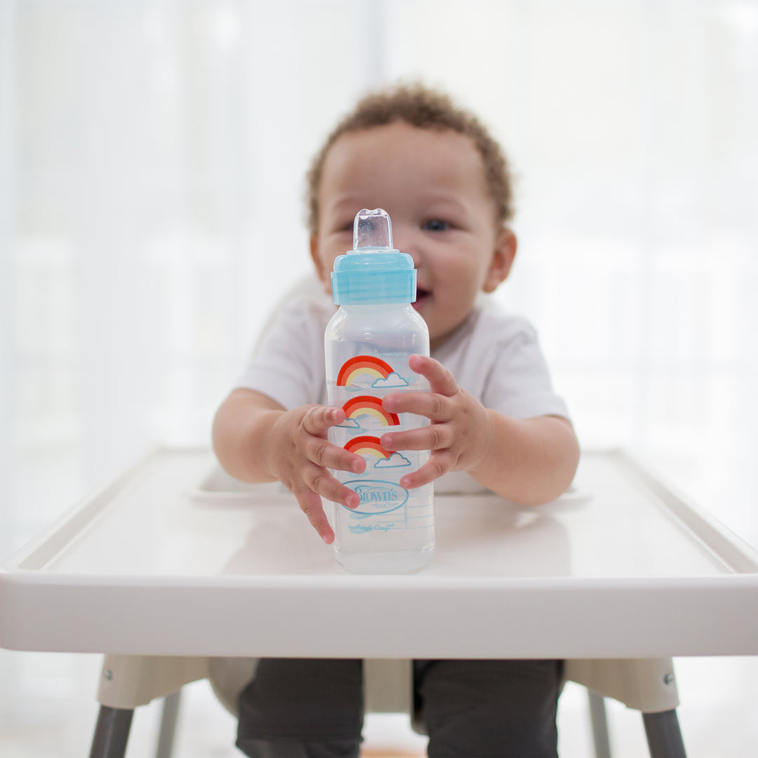 A baby sitting in a high chair holds Dr. Brown's Natural Flow® Anti-Colic Options+™ Narrow Sippy Bottle Starter Kit, 8oz/250mL. The bottle is decorated with a cloud and rainbow design. The softly blurred background emphasizes the bright and cheerful atmosphere.