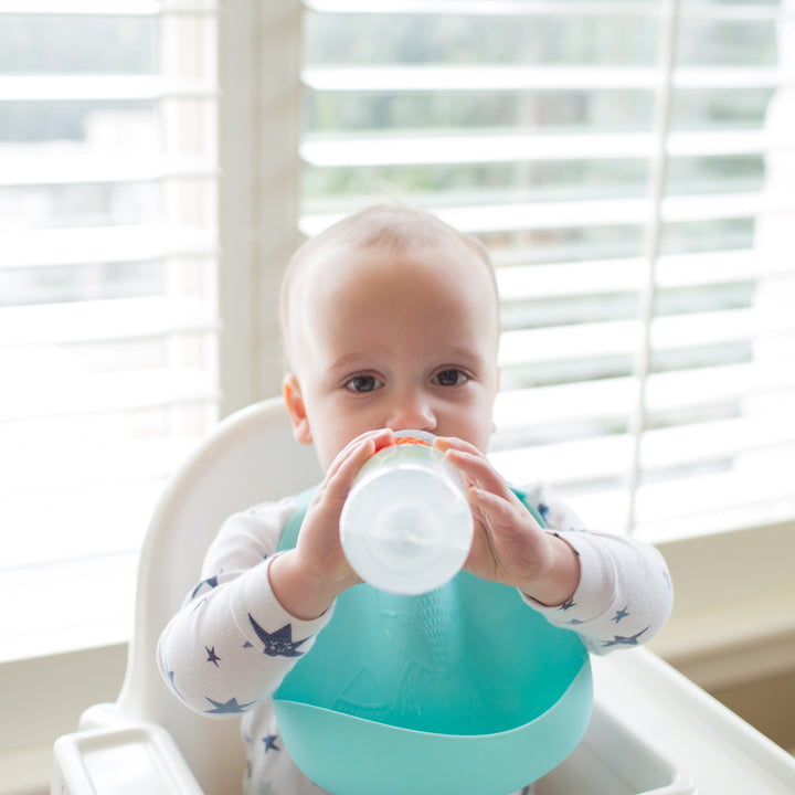 A baby in a high chair by a window with white blinds, dressed in an outfit adorned with star patterns, sips from a Dr. Brown's® Milestones™ Narrow Sippy Bottle.