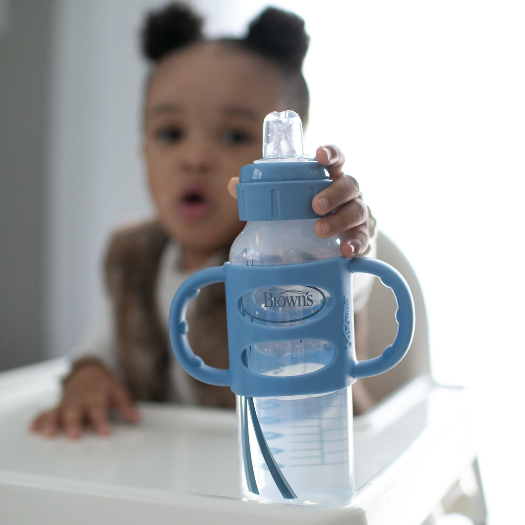 A toddler sits in a high chair, holding a Dr. Brown’s® Milestones™ Narrow Sippy Spout Bottle with Silicone Handles, 8 oz/250 mL. The child is slightly blurred in the background while the focus remains on the bottle in the foreground. The setting is indoors, suggesting a cozy home environment.