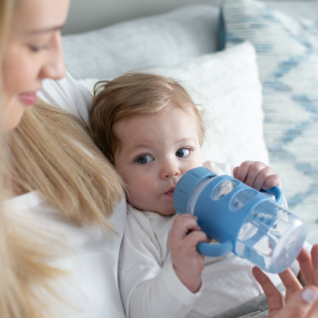 A baby with brown hair is drinking from a Dr. Brown's® Milestones™ Wide-Neck Sippy Spout Bottle with Silicone Handles while being held by a woman with long blonde hair. The woman is smiling and sitting on a light-colored sofa adorned with blue and white patterned cushions in the background.