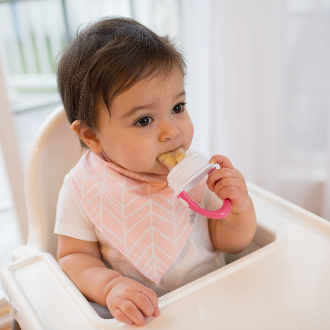 A baby sits in a high chair, joyfully munching on a snack with Dr. Brown's™ Fresh Firsts™ Silicone Feeder. Wearing a pink bib, the little one grips the pink-handled baby feeder filled with tasty treats. The softly lit interior adds warmth to the playful scene.