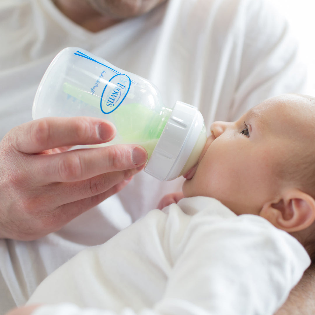 A person wearing a white shirt is feeding an infant using Dr. Brown’s Natural Flow® Wide-Neck Baby Bottle Silicone Nipple by Dr. Brown's. The bottle contains a light green liquid, while the baby, dressed in white and cradled in their arm, gazes curiously at the bottle.