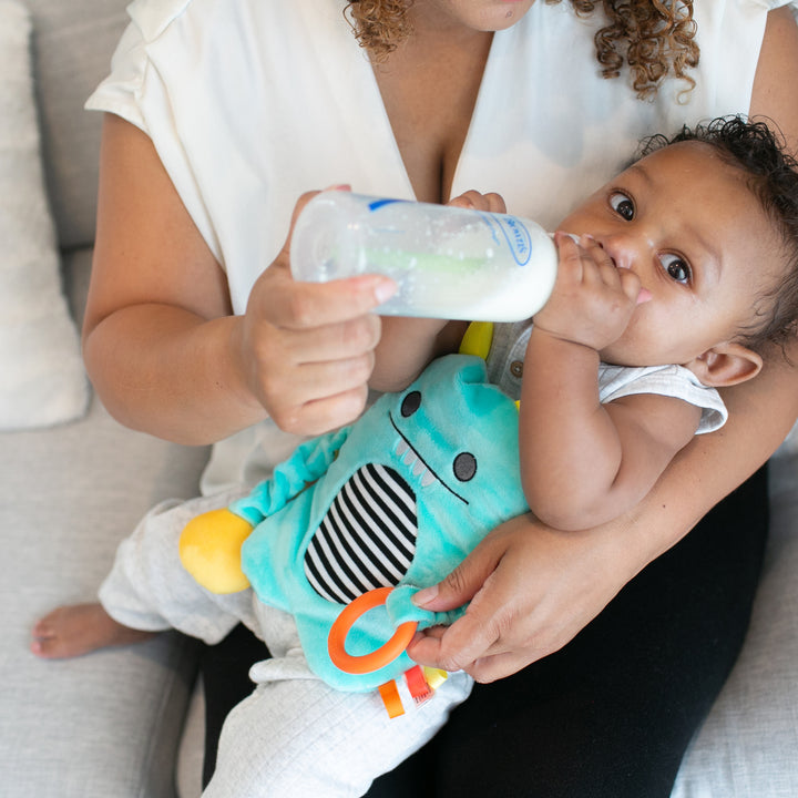 An infant in a turquoise outfit with a playful monster design is being held by an adult with curly hair on a couch, drinking from a baby bottle. The adult, wearing a white shirt, gently uses the Dr. Brown’s™ Gripebelt™ Colic Swaddling Belt for natural gas relief to ensure baby colic relief.