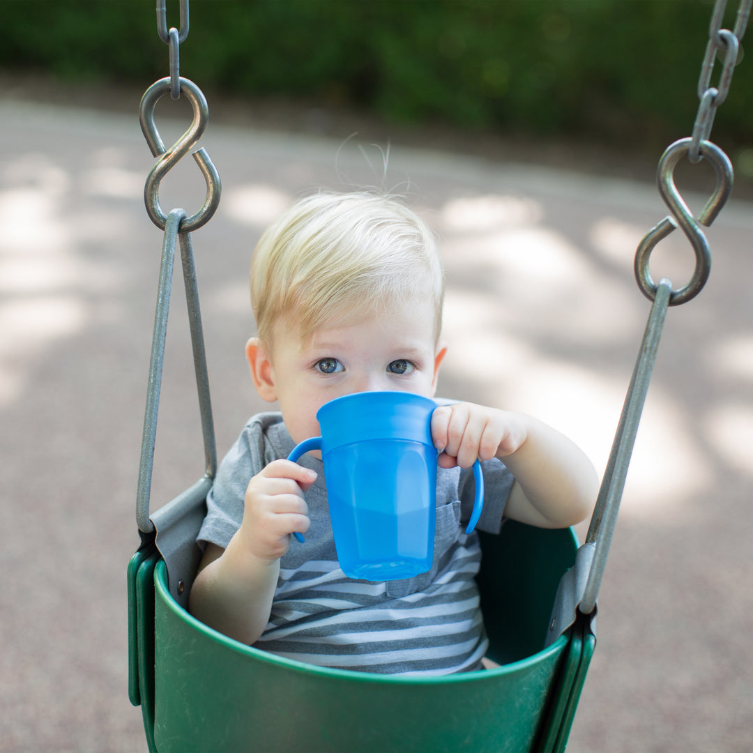 A toddler with blonde hair is sitting in a green swing, sipping from a Dr. Brown’s® Milestones™ Cheers360™ Cup with Handles, 7 oz/200 mL. The child is wearing a gray and white striped shirt, and the blurred background reveals the surface of a playground.