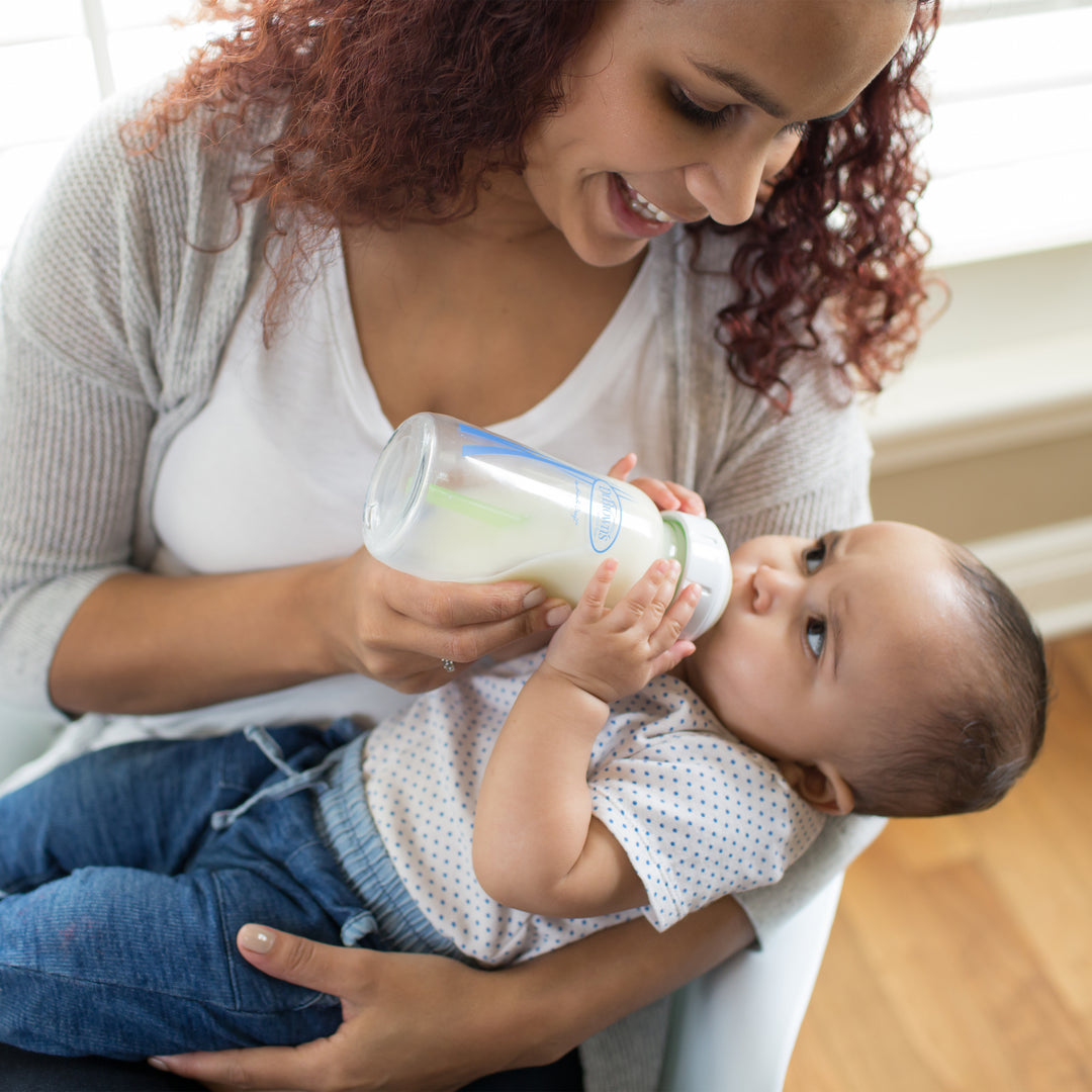 A person with curly hair is smiling warmly while feeding a baby using Dr. Brown’s Natural Flow® Wide-Neck Baby Bottle Silicone Nipple. The baby, dressed in a polka-dot shirt and jeans, looks up at the feeder, enjoying the anti-colic benefits of their wide-neck bottle in the brightly lit room.