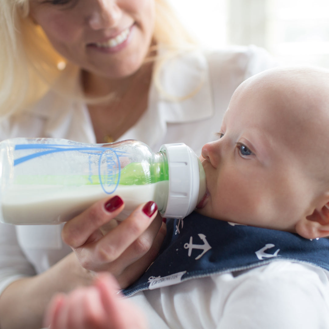 A person feeds their baby with a Dr. Brown’s Natural Flow® Anti-Colic Options+™ Wide-Neck Glass Baby Bottle, equipped with a Level 1 Slow Flow Nipple. The baby, wearing a navy blue bib adorned with white anchors, contentedly drinks milk while being held.