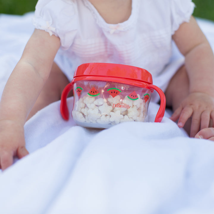 A baby in a white outfit sits on a white blanket outdoors, reaching towards Dr. Brown's Designed to Nourish Snack Cup, which features watermelon designs and a red lid, filled with small toddler snacks.