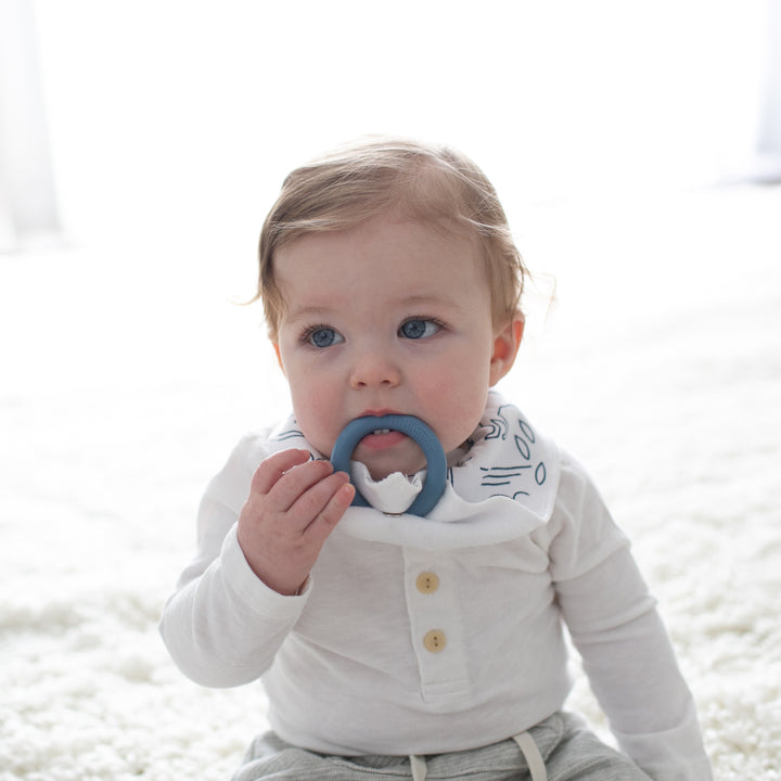 A teething baby with light brown hair is seated on a soft white surface, dressed in a white shirt and a Dr. Brown’s Bandana Bib featuring a patterned design. The baby holds the removable blue teething ring from Dr. Brown's collection and gazes slightly to the side.