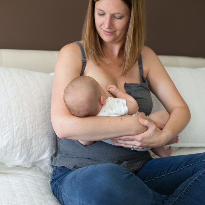 A woman with brown hair is sitting on a bed, breastfeeding an infant. She is wearing a gray tank top and blue jeans, while the baby is in a white onesie. Using Dr. Brown's ultra-absorbent Washable Breast Pads for leakage prevention, the cozy bedroom is adorned with white pillows against a brown wall.