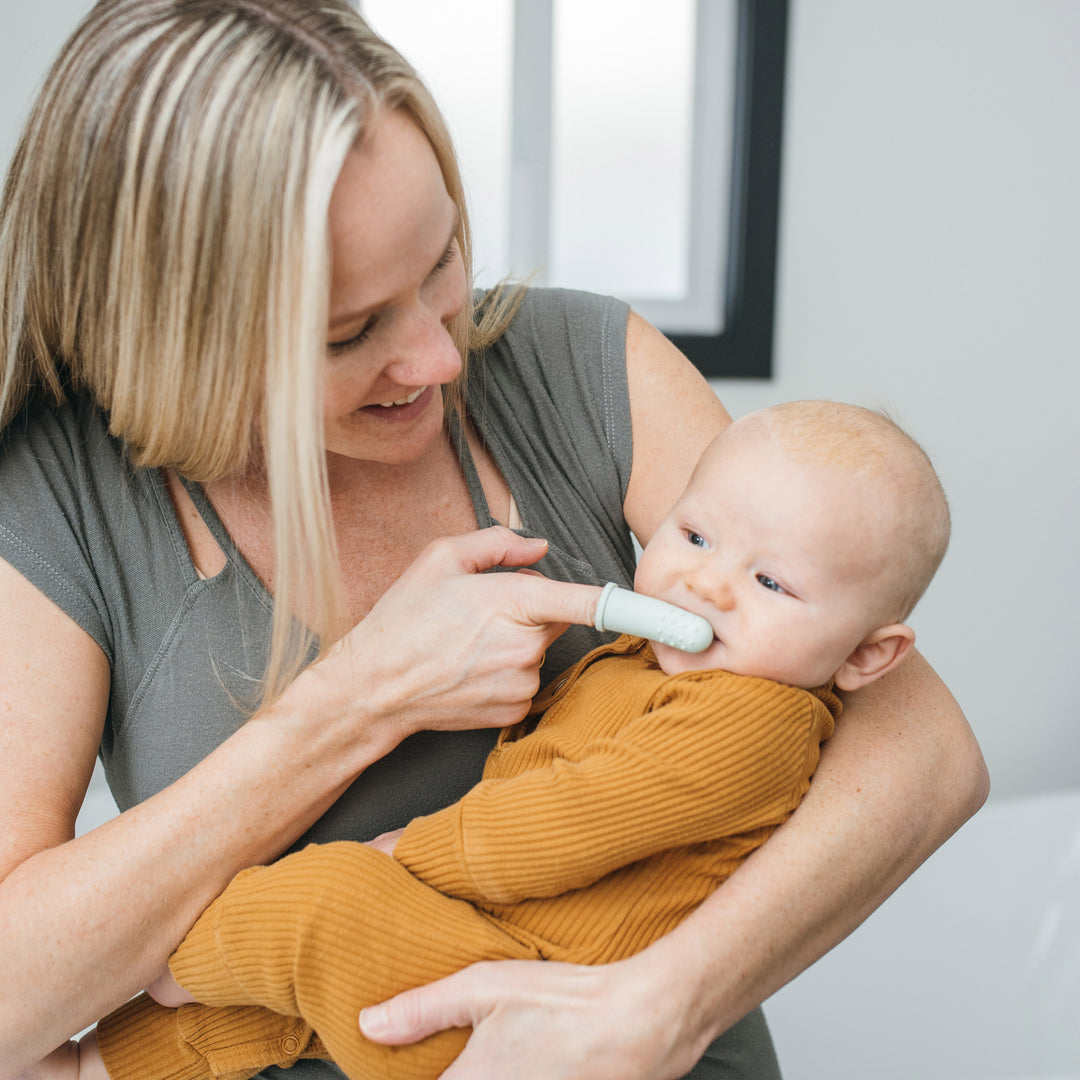 In a bright room, a woman warmly smiles at the baby held in her arms, who is dressed in a brown outfit. She gently cleans the baby's mouth using Dr. Brown's™ Silicone Finger Toothbrush with soft silicone bristles, ensuring impeccable oral hygiene.