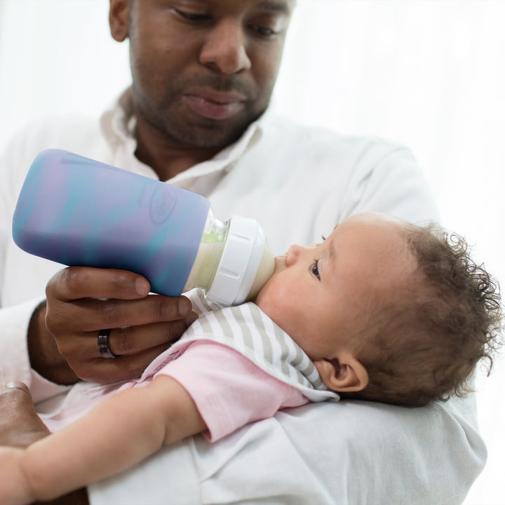 A person wearing a white shirt feeds a baby with Dr. Brown's Natural Flow® Options+™ Wide-Neck Glass Bottle covered in silicone sleeves. The baby, wearing a pink outfit and a striped bib, gazes upward while being gently held. The softly blurred background enhances the serene atmosphere of the moment.