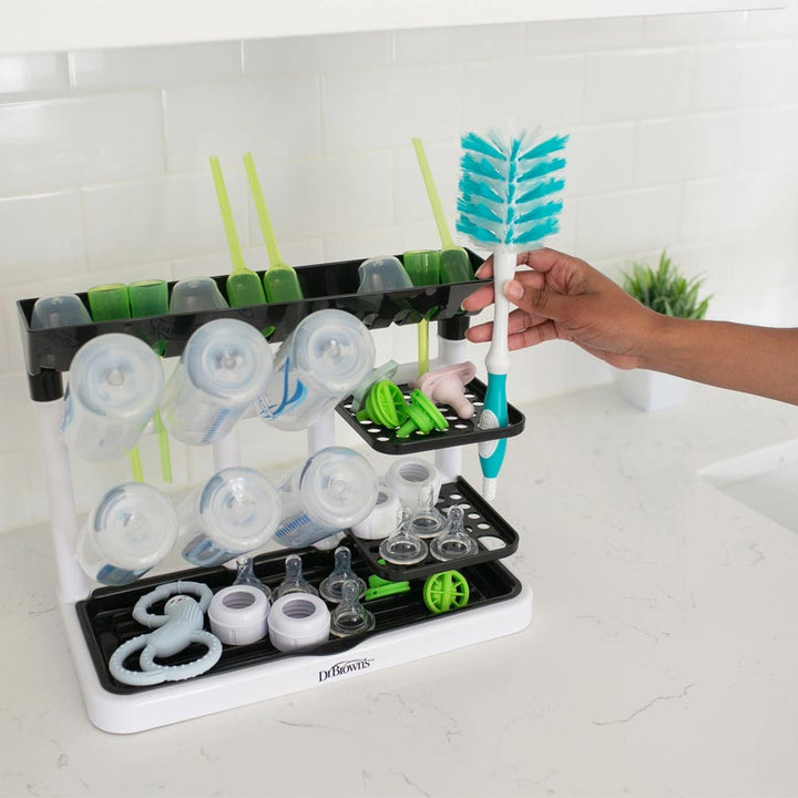 A hand places a bottle brush into the Dr. Brown’s™ Drying Tower, which is filled with various baby bottle parts such as nipples, pacifiers, and lids. This practical countertop space saver from Dr. Brown's is located on a white kitchen countertop with a white tile backsplash.