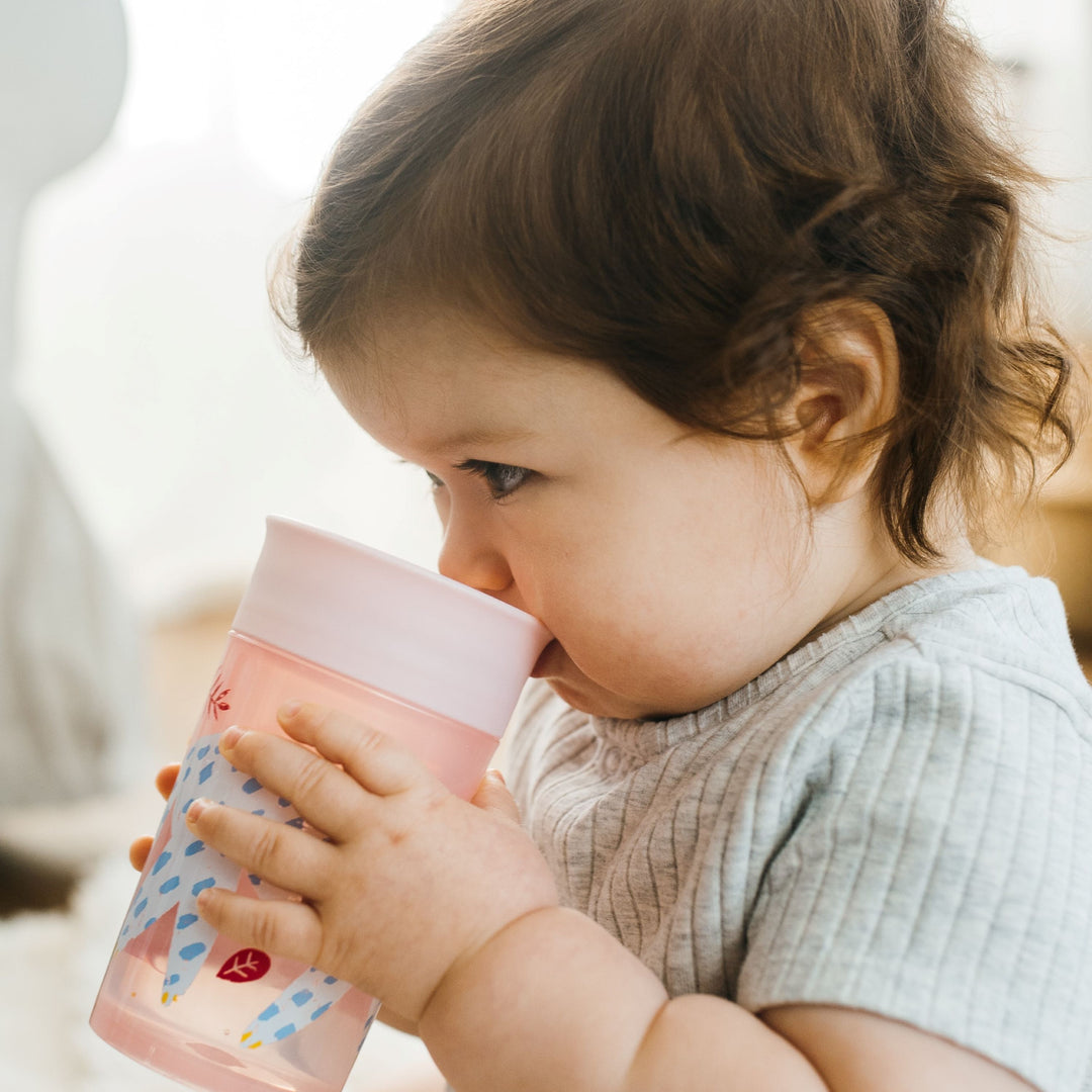 A toddler with brown hair drinks from a Dr. Brown’s® Milestones™ Cheers360™ Cup, which is pink and decorated with blue patterns. This spoutless cup features a silicone valve to prevent spills. The child, dressed in a light gray shirt, seems to be indoors against a softly blurred background.