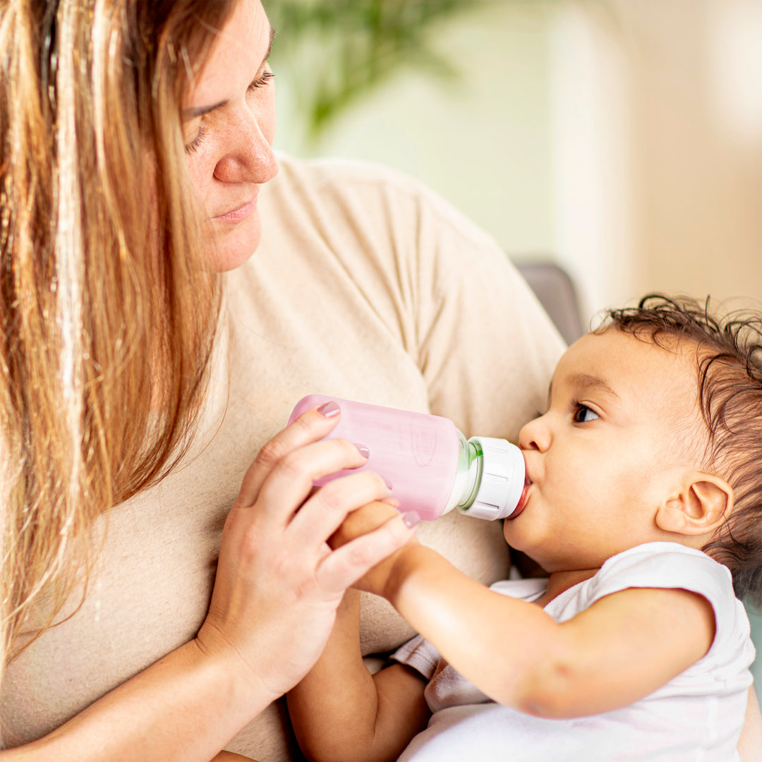 A woman with long hair holds a baby who is drinking from a Dr. Brown’s Natural Flow® Options+™ Narrow Glass Bottle Silicone Sleeves. They are seated indoors with a blurred background suggesting a cozy environment. The baby, in a white shirt, delights in their feed from one of the glass baby bottles designed for comfort and safety by Dr. Brown's.