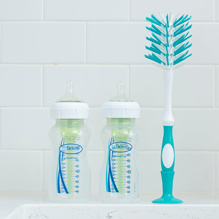 Two BPA-free Dr. Brown's baby bottles sit next to a Dr. Brown’s Natural Flow® Deluxe Bottle Brush, featuring a handy nipple cleaner, on a white countertop against pristine white tiled walls.