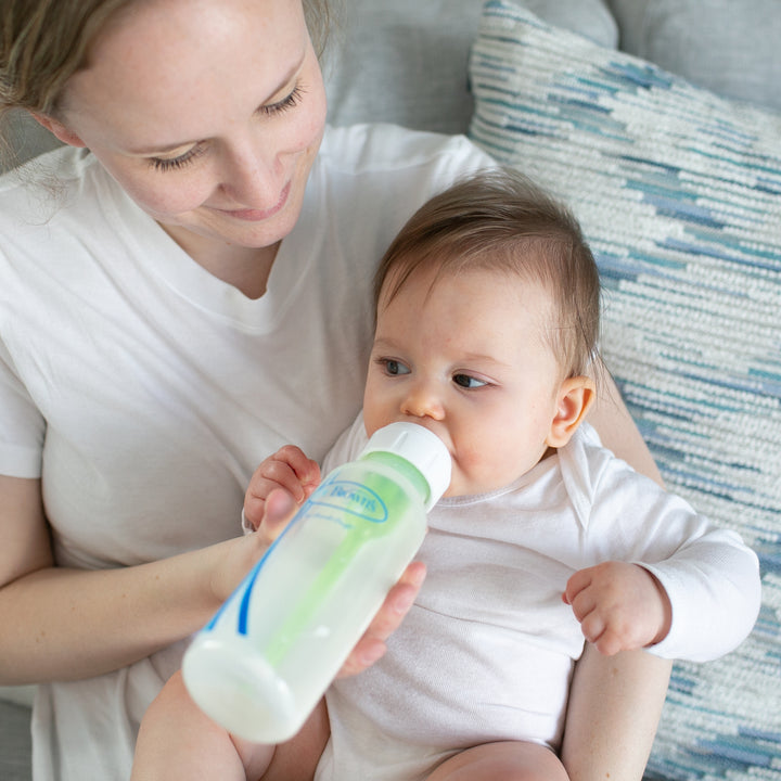 A woman sits on a couch, gently cradling a baby who’s peacefully drinking from a Dr. Brown's Natural Flow® Anti-Colic Options+™ Narrow Baby Bottle, featuring a Level 1 Slow Flow Nipple. Both are dressed in white clothing, with a patterned blue and white pillow in the background.