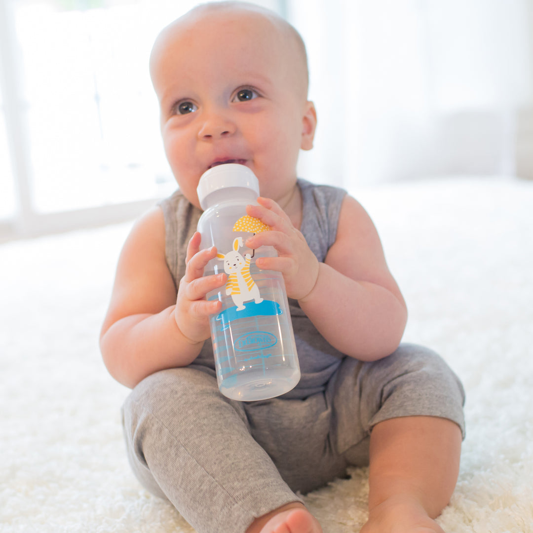 A baby in a gray outfit is sitting on a fluffy white surface, happily chewing on Dr. Brown's® Milestones™ Narrow Sippy Bottle, which features a cartoon animal design and a soft silicone sippy spout, making it ideal for transitioning from bottle to cup. Light streams in from a window in the background.